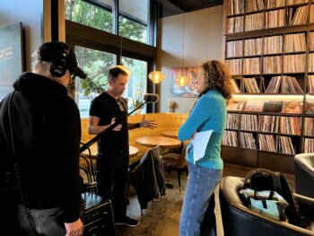 Andrea interviewing a guest, with the audio engineer and microphone boom nearby in front of a large street facing window and a wall of vinyl records in the background.